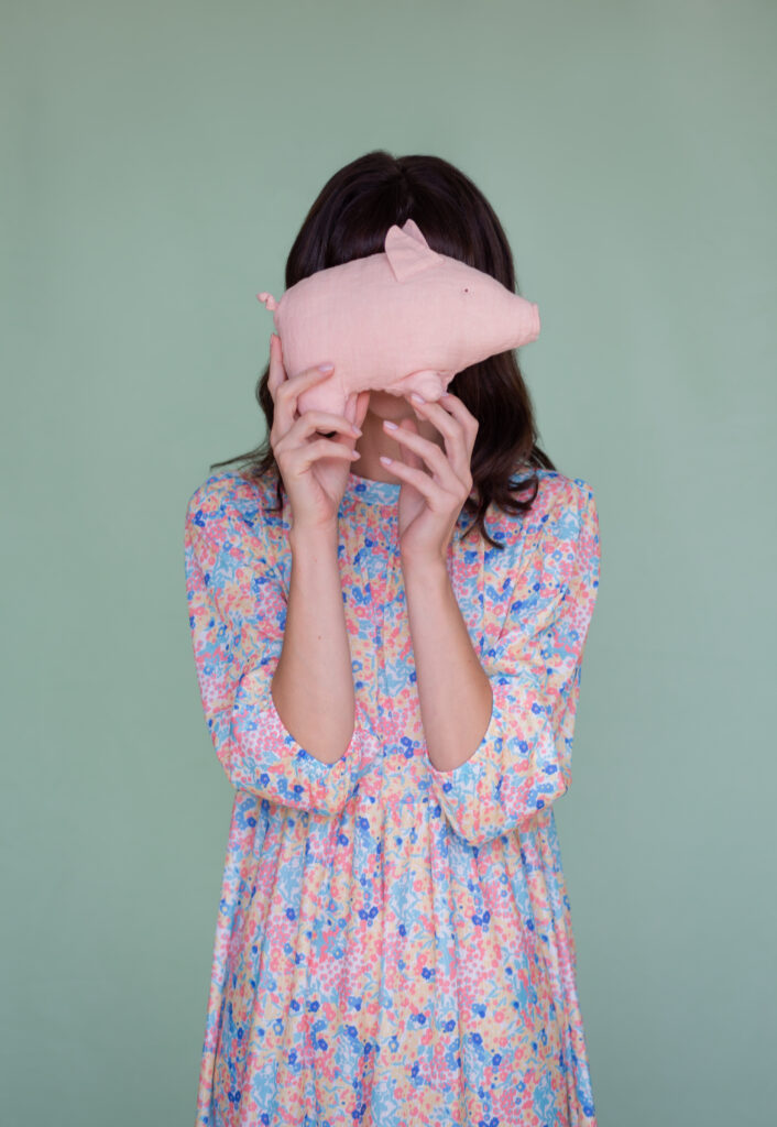 Eine Frau in einem farbenfrohen, geblümten Kleid hält sich spielerisch ein rosa Stoffschweinchen vor das Gesicht. Hellgrüner Hintergrund.